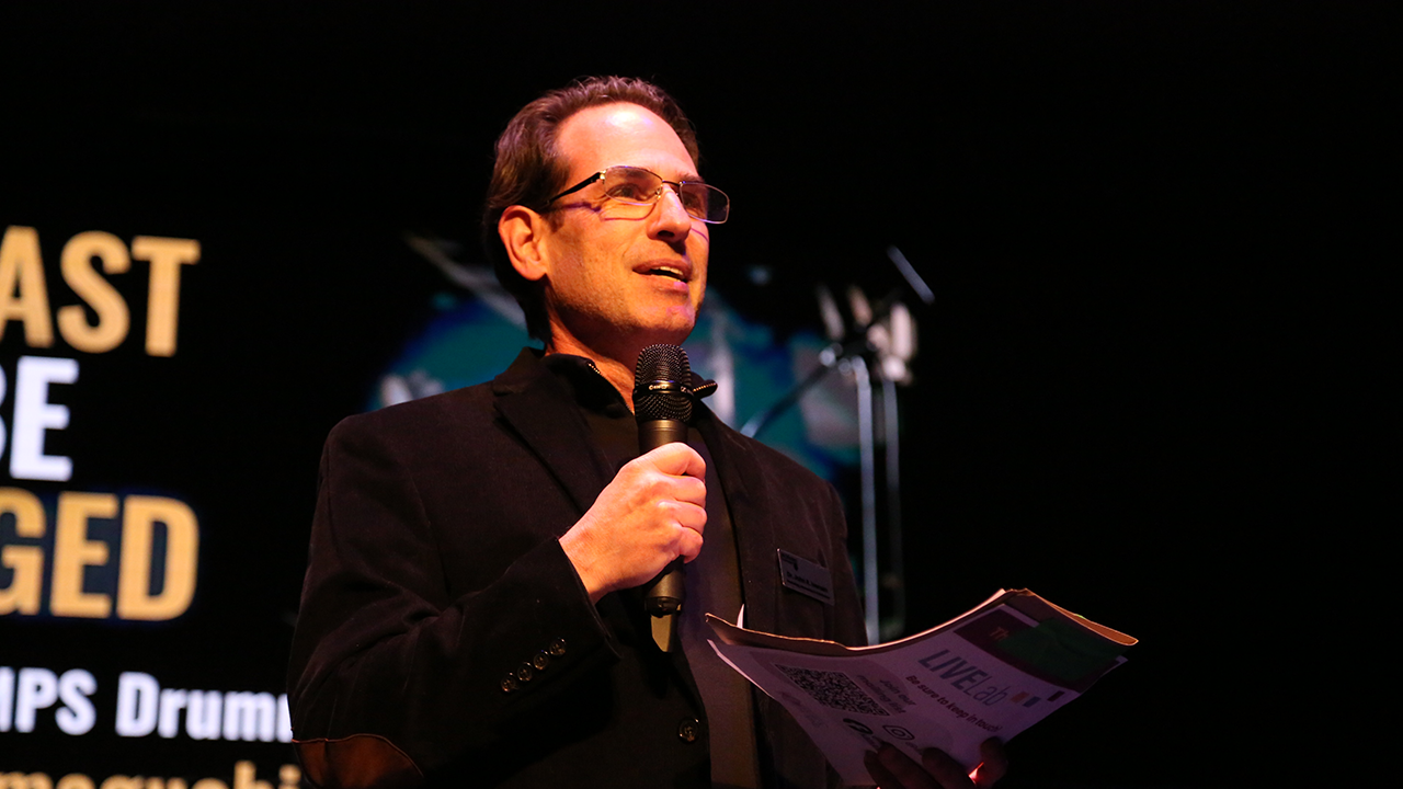 Man in a dark blazer speaks into a handheld microphone on a stage, holding a program flyer.