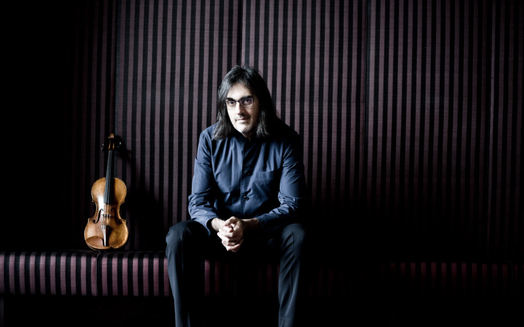 Portrait of a man with shoulder-length dark hair and glasses, in a blue shirt, sitting in a dim room with a violin resting beside him on striped burgundy walls.