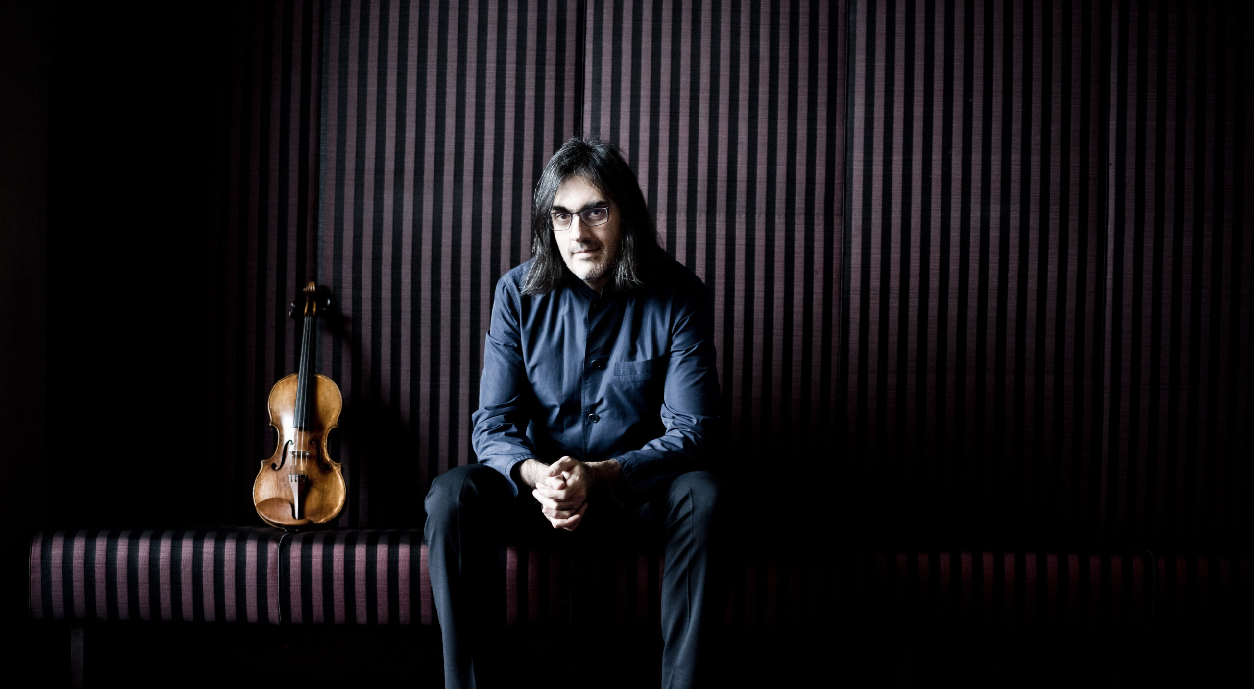 Portrait of a man with shoulder-length dark hair and glasses, in a blue shirt, sitting in a dim room with a violin resting beside him on striped burgundy walls.