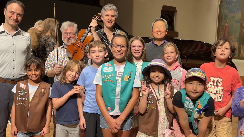Group of smiling children in Scout vests posing with adult musicians on a stage; a violin and a grand piano are in the background.