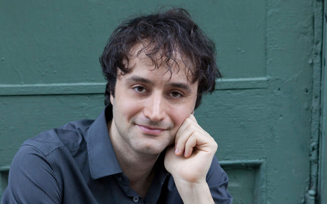 Man with dark hair wearing a blue shirt rests his chin on his hand and smiles softly, in front of a green door background.
