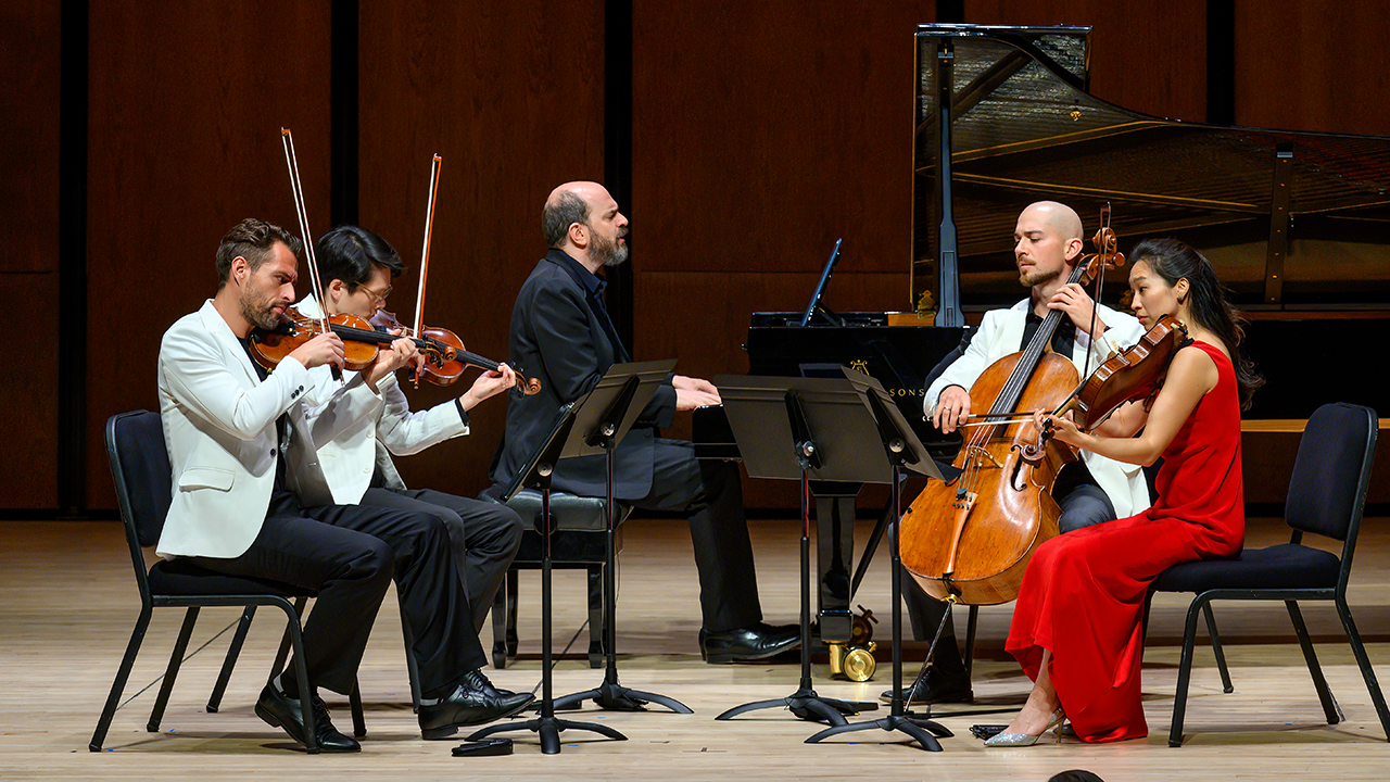 Chamber ensemble performing on stage: two violinists, a pianist, a cellist, and a violinist in a red dress.
