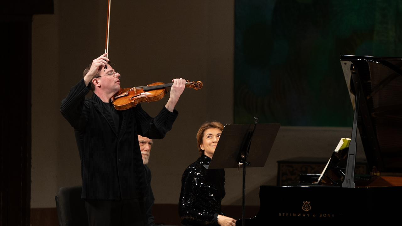 Violinist raises bow while playing on a concert stage; a pianist sits at a Steinway & Sons grand piano nearby.