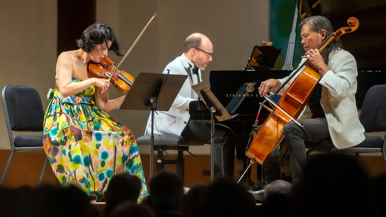 Violinist in a vibrant floral dress plays violin on a stage beside a pianist and a cellist; audience silhouettes in foreground.