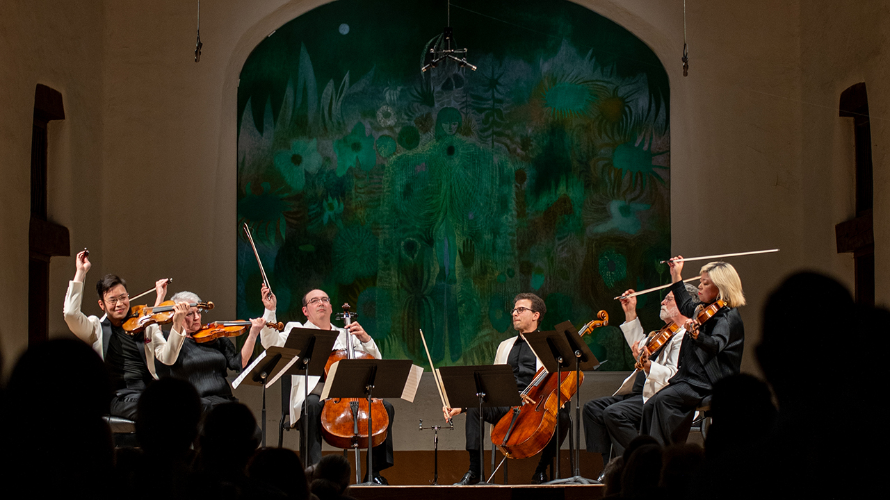 Orchestra of string players performing on a stage with a green abstract mural backdrop and audience silhouettes in the foreground.