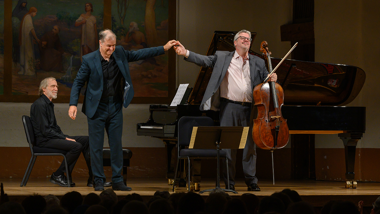 Three male musicians bow on a concert stage: a pianist seated at a grand piano, and two standing with joined hands after a performance; audience in foreground.