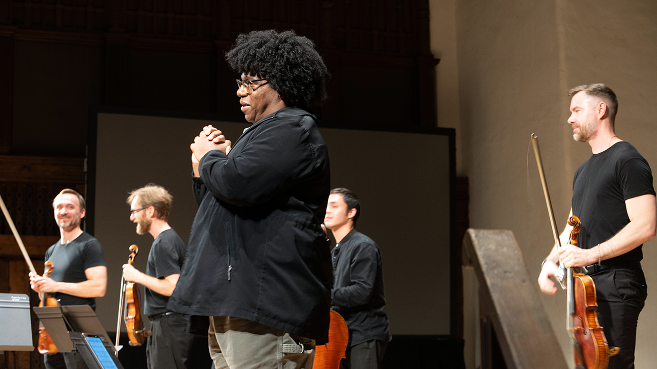 A performer with a large afro claps at the front while a string ensemble stands behind with violins and a cello on stage.