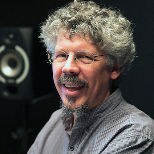 Smiling man with curly gray hair, glasses, and a goatee in a recording studio near a speaker.