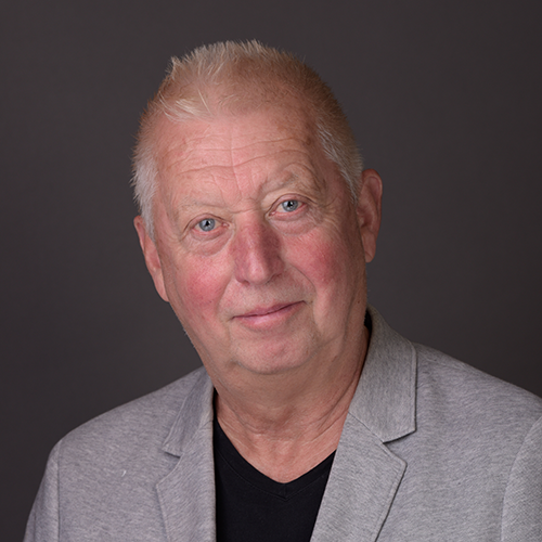 Portrait of an older man with short gray hair wearing a light gray blazer over a black shirt, looking at the camera in a neutral studio setting.