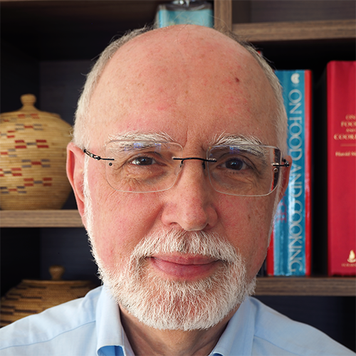 Close-up portrait of an older man with white beard and glasses, standing in front of a bookshelf.